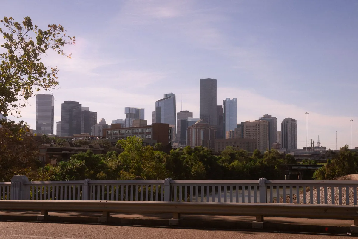The downtown Houston skyline of glass and stone towers seen from a road bridge, with the bridge balustrade and steel guardrail across the foreground and a band of trees between