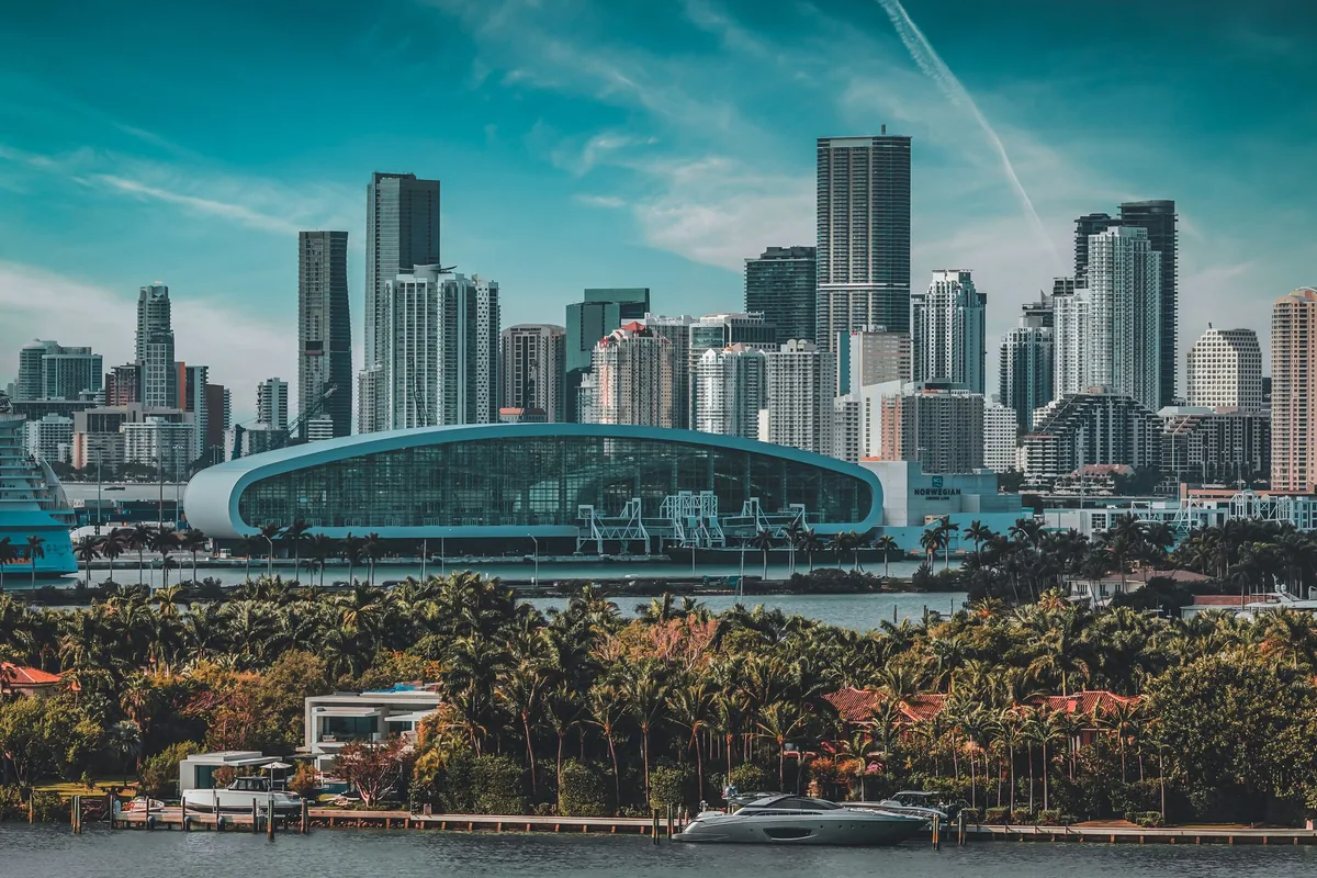 The downtown Miami skyline seen across Biscayne Bay, with the curved glass roof of a cruise terminal in front of the towers and palm-lined waterfront houses and a moored yacht in the foreground