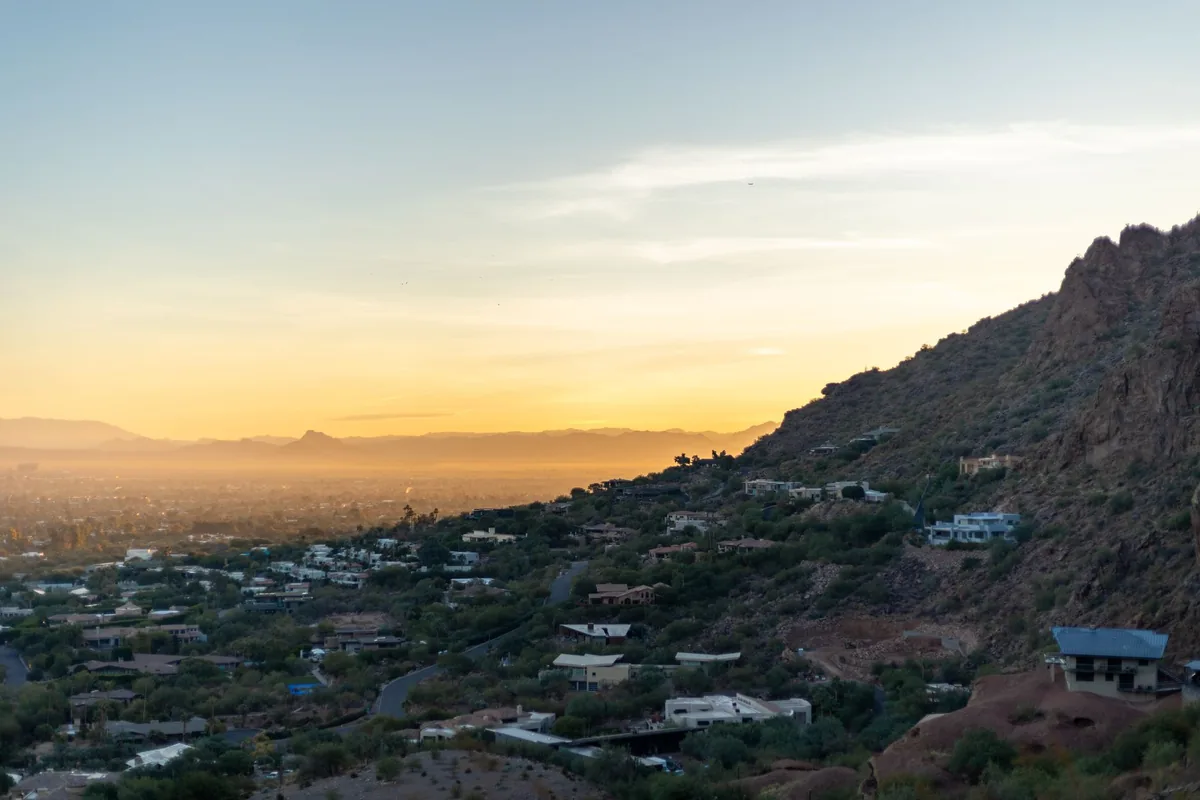 Sunset over the Phoenix valley seen from high on a desert mountainside, houses set among scrub along the slope on the right and the flat city spreading to distant ranges on the left